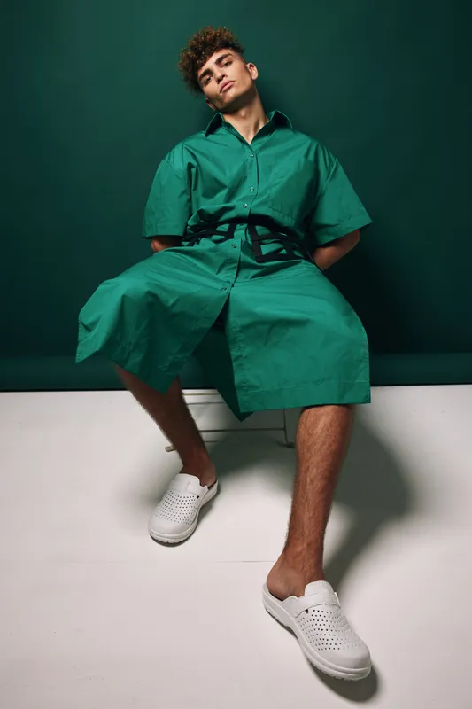 Young man wearing green oversized shirt and shorts sitting on white bench with relaxed pose against dark green background in studio. Casual fashion and modern style with white slip on shoes.