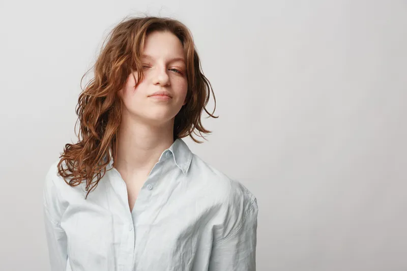 Young woman with curly hair wearing white shirt poses with confident expression on light background. Concept of natural beauty, casual style, and self-confidence.