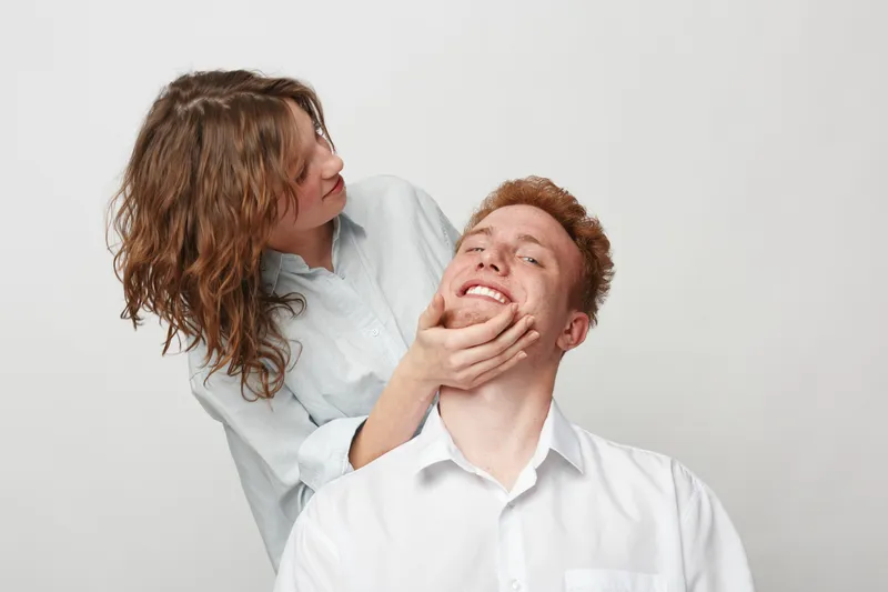 Young woman gently holds smiling man’s face in white shirts against plain background. Concept of affection, playful interaction, friendship, and positive emotions between people.