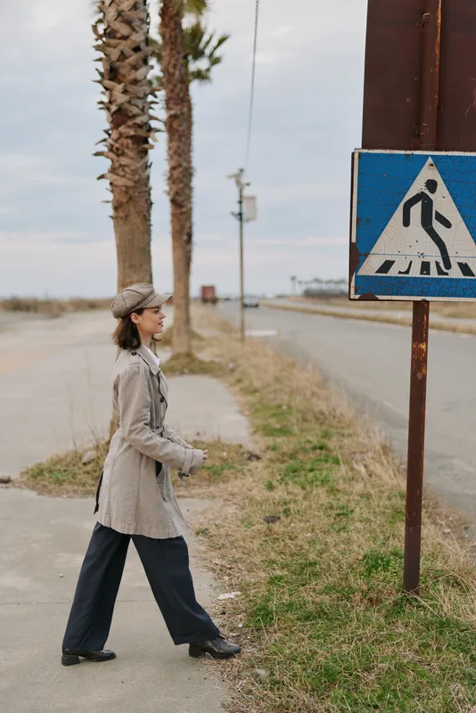 Woman in coat and hat walking on sidewalk near pedestrian crossing sign on quiet street with palm trees and cloudy sky. Casual urban lifestyle and outdoor concept.