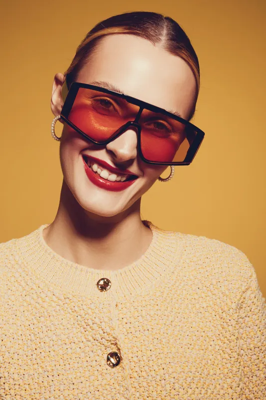 Smiling woman with red lipstick wearing oversized orange sunglasses and yellow knitted sweater on yellow background. Concept of fashion, style, confidence, and modern accessories.