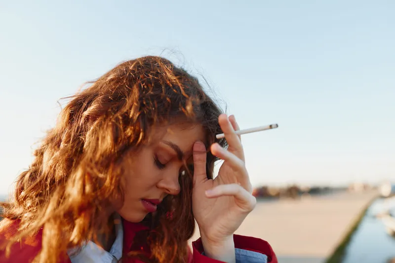 Young woman with curly hair holding cigarette near face outdoors, thoughtful expression, soft natural light, blurred background, casual clothing, urban setting