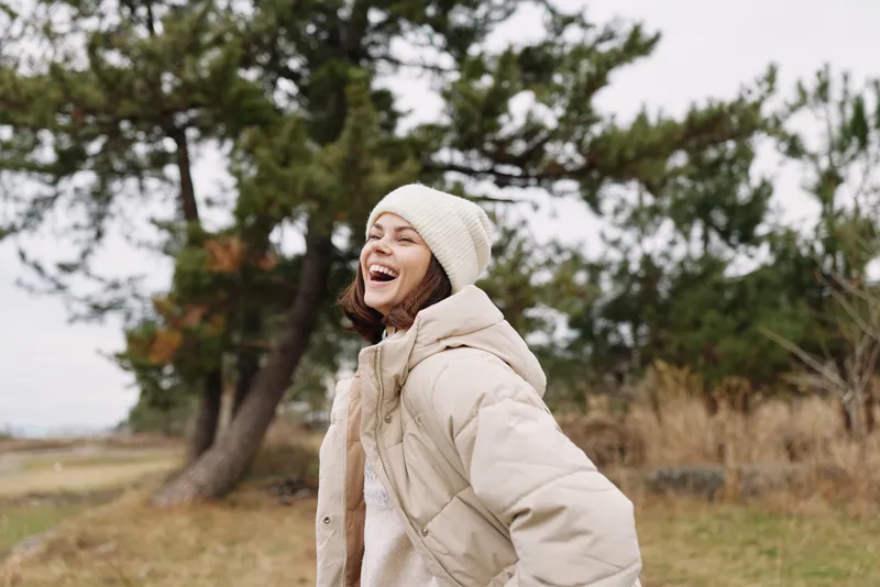 Woman outdoors wearing a beige coat and beanie, smiling joyfully on a windy autumn day with trees in the background, showcasing warmth, comfort and candid outdoor fashion.