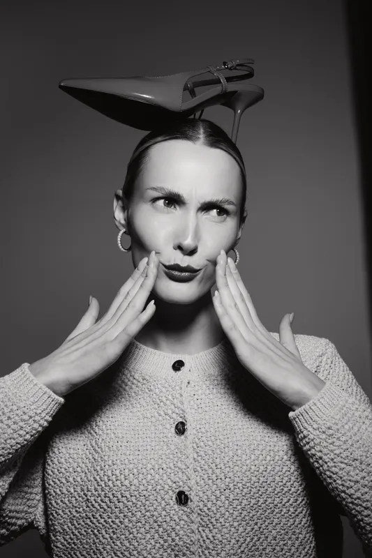 Caucasian woman with fair skin and vintage hairstyle touches her face, wearing a high heel on her head in black and white image.