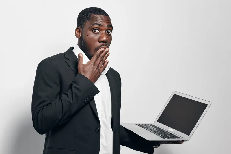 African American man with dark skin in black suit holding laptop with blank screen and covering mouth with hand in surprise or shock on white background.