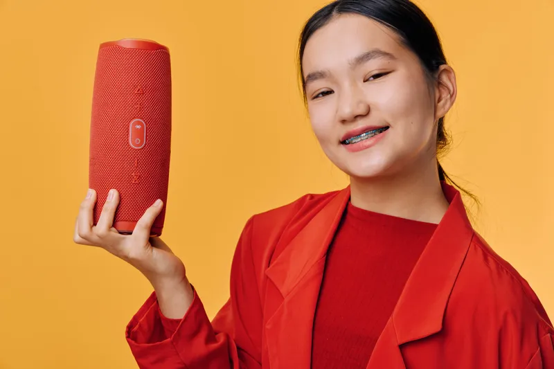Young woman holding red wireless speaker smiling with braces wearing red jacket on orange background for technology and music concept. Portrait, gadget, device and entertainment.