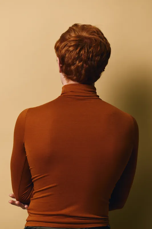 Man with red hair wearing brown turtleneck sweater stands with back to camera against beige background in studio or indoor setting.