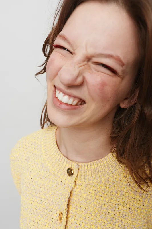 Close-up of a young woman with brown hair smiling widely and squinting eyes wearing a yellow knitted sweater. Bright studio background highlights natural expression and joyful mood.