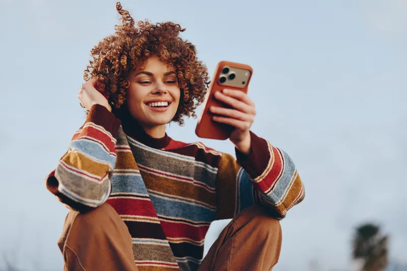Woman lifestyle outdoors, smiling selfie with smartphone, curly hair, casual striped sweater, close portrait, bright daylight, natural candid moment, phone in hands for social media sharing