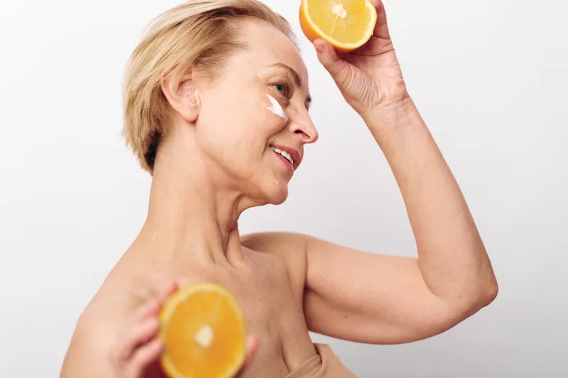 Mature woman with short blonde hair applies natural face cream and holds two orange halves near her face. Concept of skincare, healthy lifestyle, and natural beauty care.