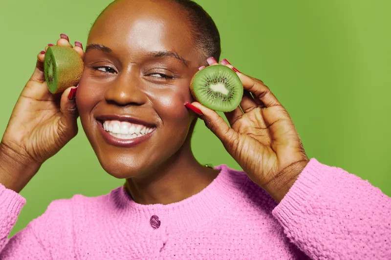 Smiling black woman with dark skin holds two halves of kiwi fruit near her face wearing pink sweater. Concept of healthy lifestyle, natural food, and joyful mood on green background.