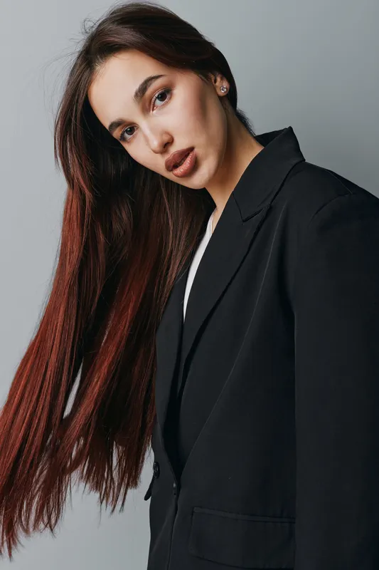 Young woman with long brown hair wearing black blazer posing against gray background in studio. Portrait of confident female model with natural makeup and serious expression looking at camera.
