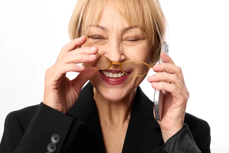 Cheerful mature woman smiling and laughing with eyes closed, holding eyeglasses by ear during phone call, business suit, studio portrait on white background