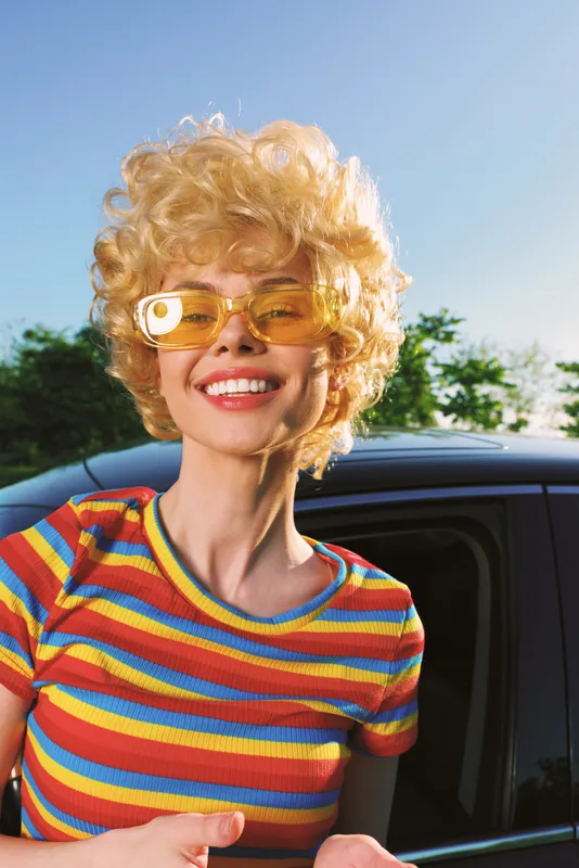 Cheerful woman outdoors, smiling with sunlit look, vibrant striped shirt, retro vibe, casual pose, warm colors, blue sky, lush greenery, bright moment, carefree mood, natural setting