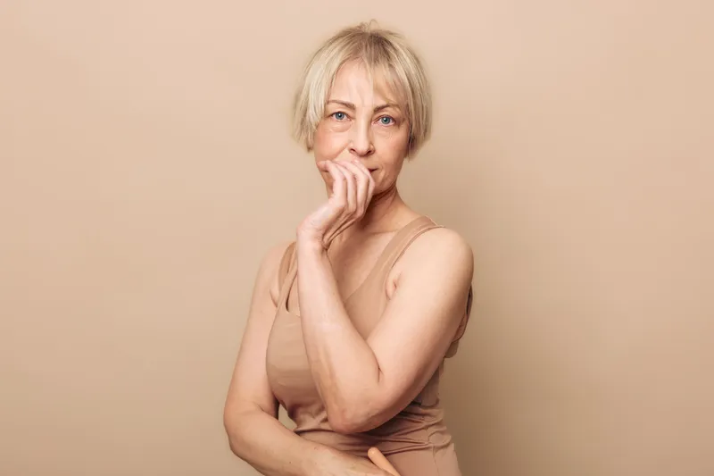 Thoughtful middle aged woman with short blonde hair poses with hand near face wearing beige sleeveless top. Neutral background creates calm mood concept for confidence and contemplation.