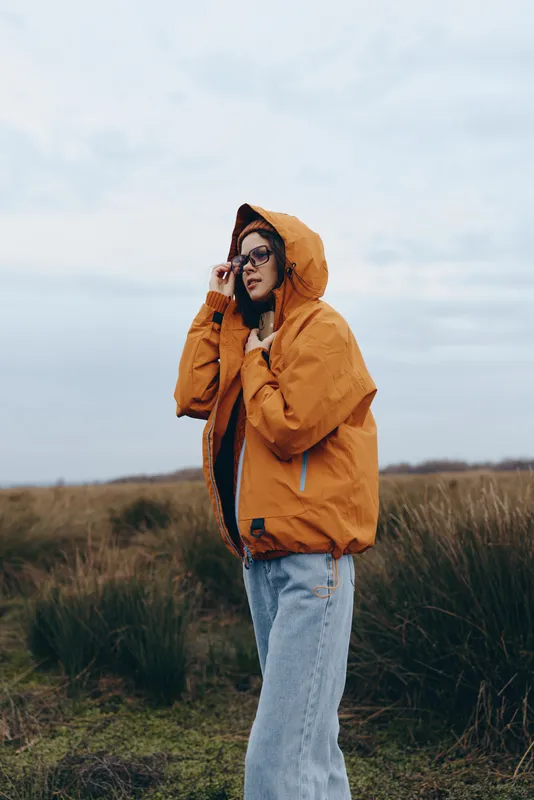 Woman lifestyle portrait outdoors in autumn, wearing an orange jacket with hood, standing in a grassy field, wind moving hair and scarf, calm mood, natural light and cloudy sky