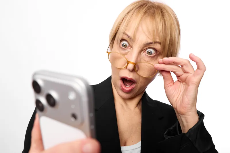 Surprised elderly business woman with glasses holds smartphone closeup, wide eyed look, office blazer, shocked gesture, isolated on neutral white background
