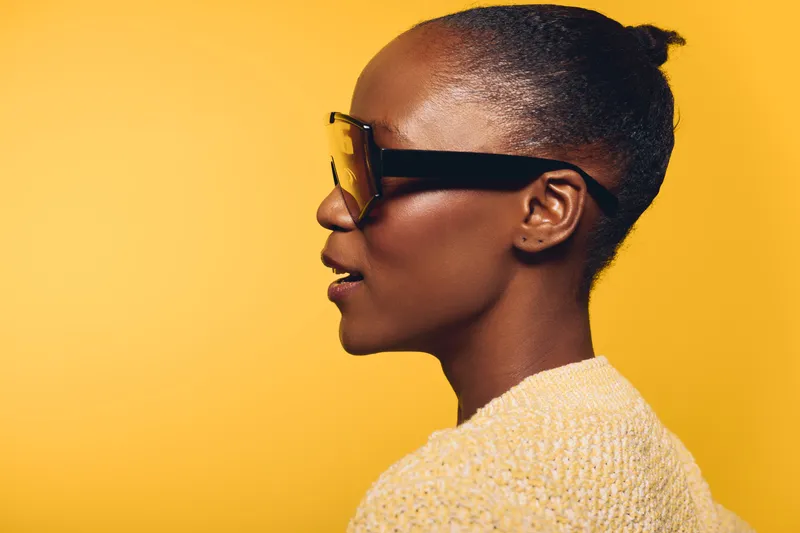 Profile of a dark skinned African woman wearing large sunglasses and a textured sweater against a yellow background. Concept of fashion, style, and modern accessories.