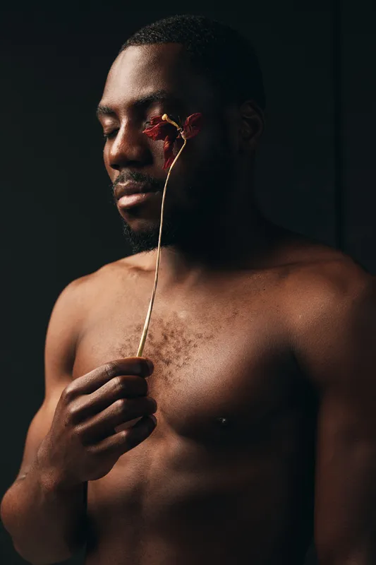 Creative conceptual portrait of African man with closed eyes holding dried flower near face in dark studio. Artistic visual style with emotional expression and bare chest.