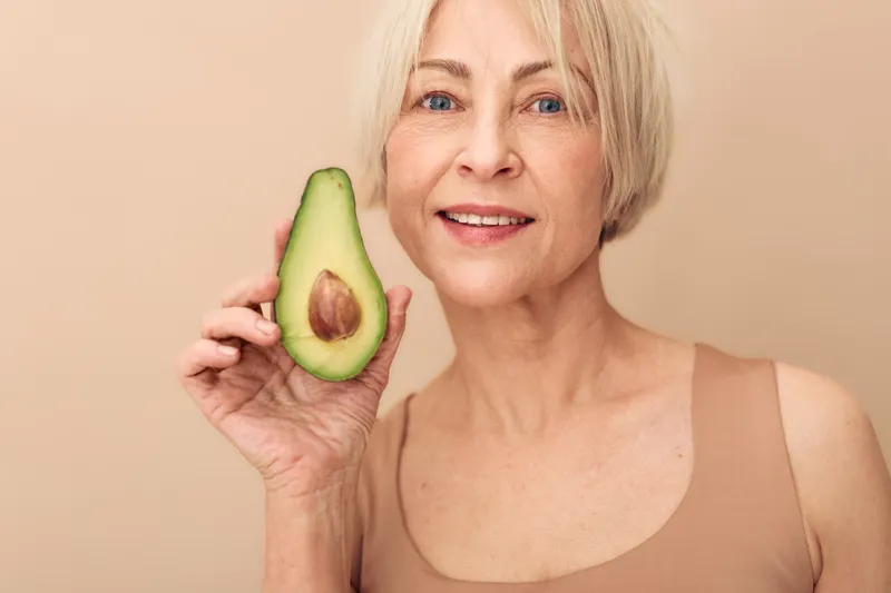 Smiling mature woman with short blonde hair holding half of avocado near face on beige background. Concept of healthy lifestyle, nutrition, and natural beauty care for seniors.