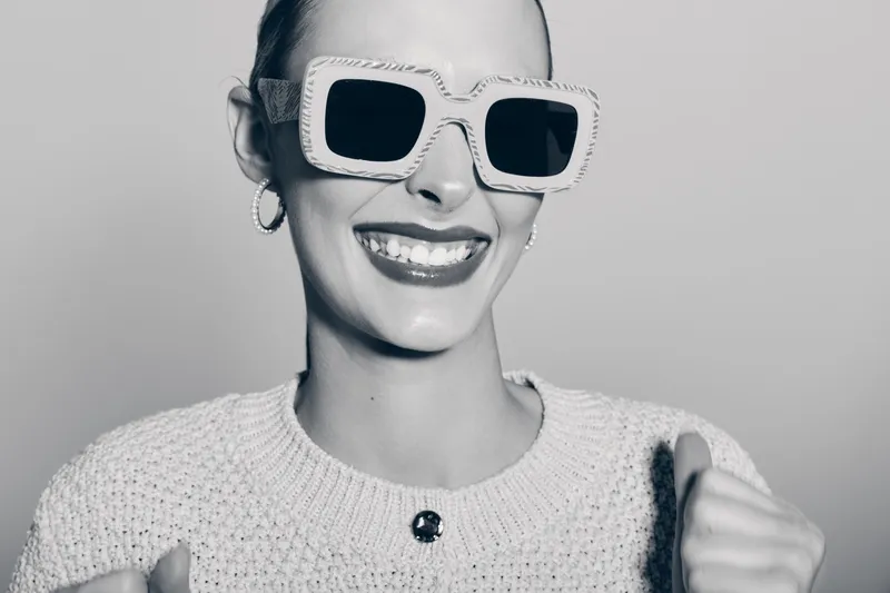 Smiling white woman with pale skin wearing large sunglasses and knitted sweater shows joyful mood in studio portrait.