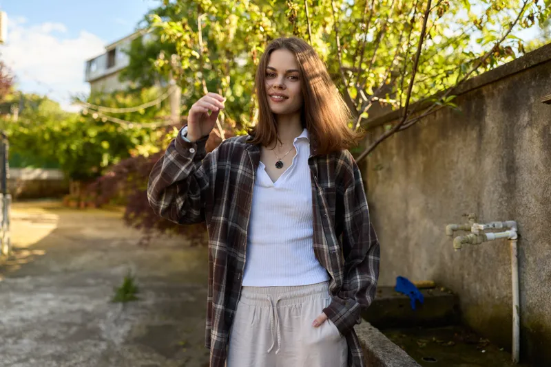 A woman in casual jacket and plaid shirt stands outdoors, smiling under sunlight on a street. A candid lifestyle moment highlighting confidence, style, and everyday fashion