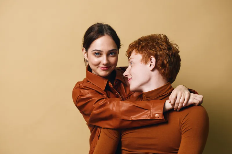 Young couple embracing indoors with warm smiles, wearing brown clothing. Woman hugs man closely, expressing affection and connection on neutral background.