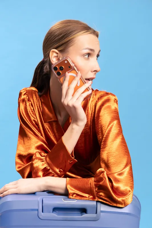 Young woman in shiny orange blouse holds smartphone to ear while leaning on blue suitcase against blue background. Concept of travel, communication, and fashion lifestyle.