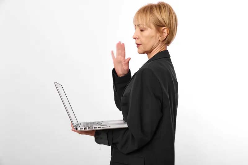 Senior woman with laptop on white background raises hand to stop, speaking in video call, focused expression, remote communication, modern technology today