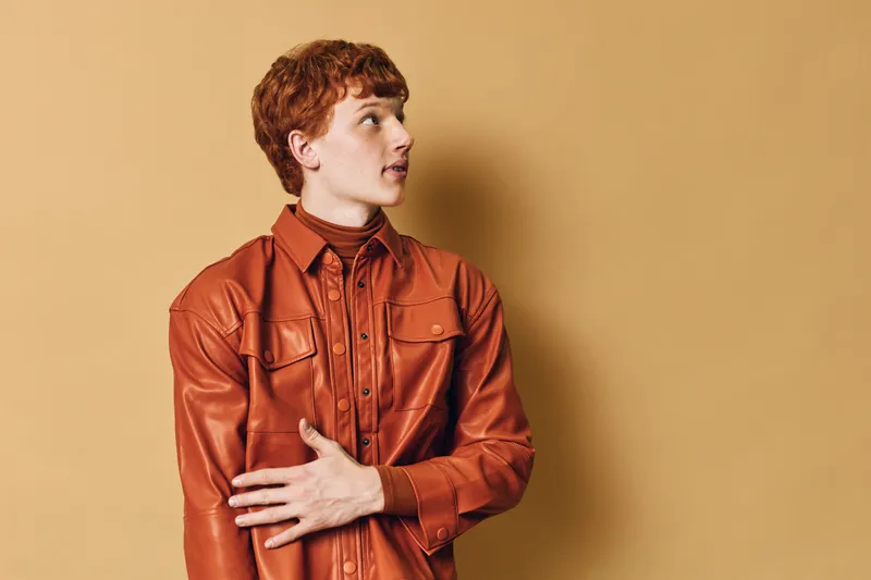 Young man with red hair wearing brown leather jacket and turtleneck looking sideways on beige background. Profile portrait with casual style and fashion expression indoors.