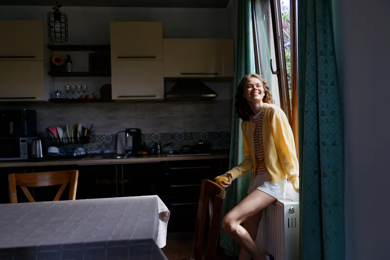 Bright morning scene of a cheerful woman in a kitchen by a sunlit window, wearing a yellow cardigan and striped top, relaxed pose, emphasizing home lifestyle and cozy everyday moments.