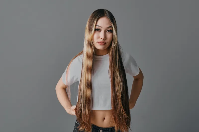 Young woman with long straight hair wearing white crop top standing with hands on hips looking at camera in studio with gray background and neutral expression.