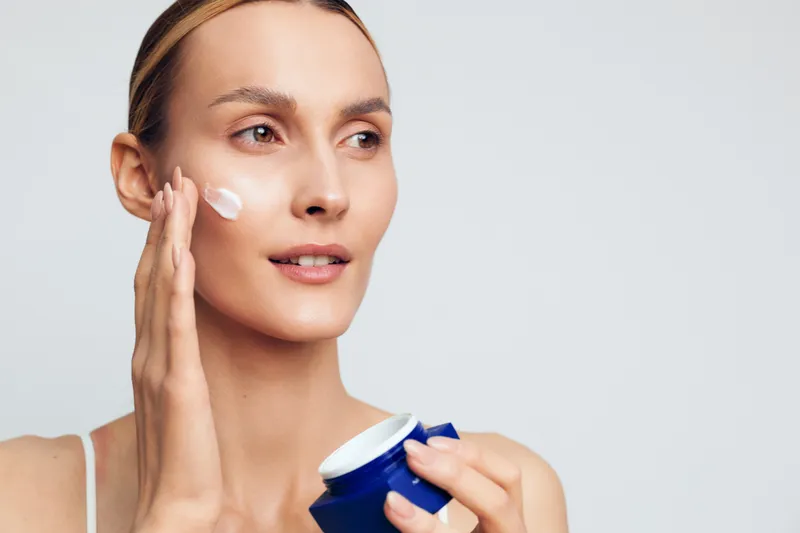 Woman skincare routine applying moisturizer cream to face cheek, holding blue jar, gentle beauty spa hygiene moment, calm expression, soft studio light, closeup natural skin