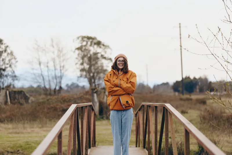 Woman lifestyle portrait, standing woman on a wooden bridge in rural nature during autumn, yellow jacket and jeans, smiling outdoors with calm mood, travel scenery