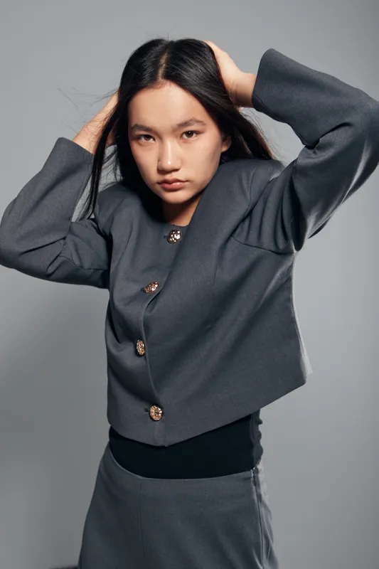 Young woman in gray blouse and skirt posing with hands in hair on gray studio background. Asian female model with long black hair and serious expression in fashion outfit.