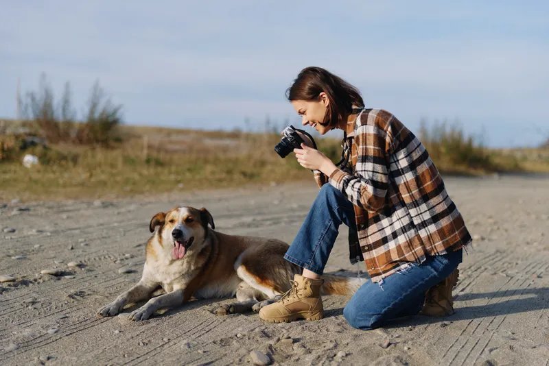 Woman kneels on a sandy landscape, camera raised to photograph a relaxed dog lying nearby, under a clear sky and wide open outdoor space in daylight.
