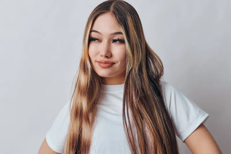 Young woman with long straight hair wearing white t shirt posing with confident smile in studio environment against light background for portrait and lifestyle.