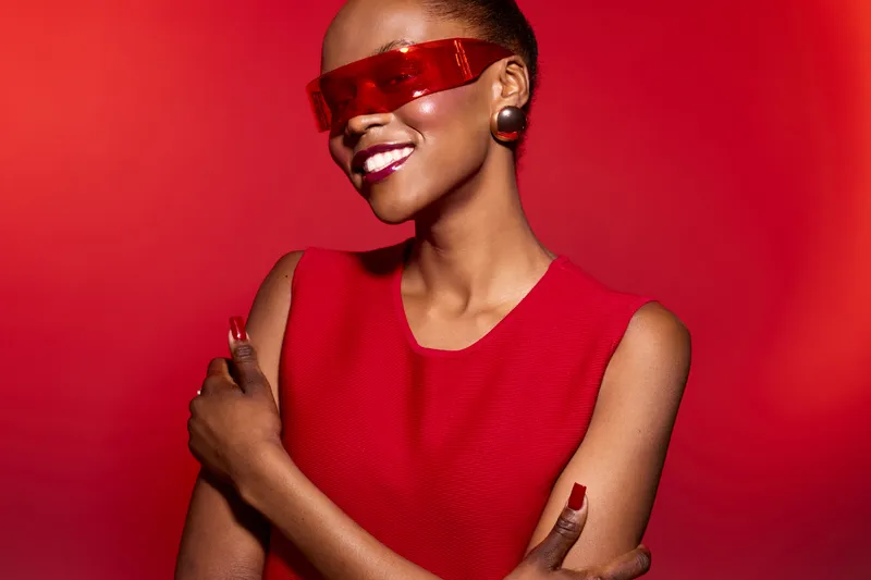 Confident African woman with dark skin wearing red sleeveless dress and futuristic red sunglasses smiles with crossed arms on red background. Concept of fashion, style, and modern beauty.