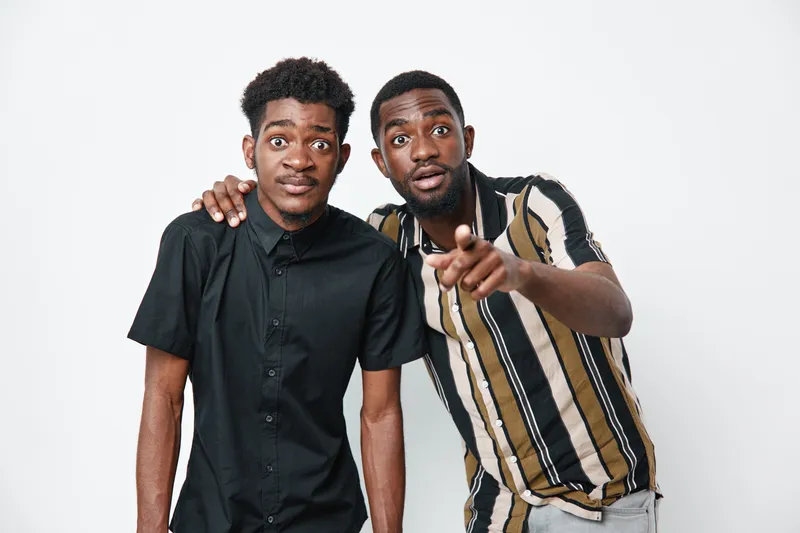 Two young black men with dark skin posing together on white background, casual clothing, one pointing forward and the other with arm on shoulder, friendship and connection.