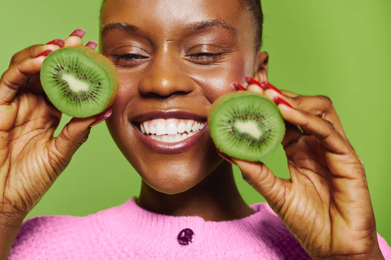 Smiling black woman with dark skin holds two kiwi halves near face wearing pink sweater on green background. Concept of healthy eating, fresh fruit, natural beauty and wellbeing.