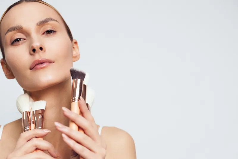 Young woman with flawless skin holds makeup brushes near face applies foundation with soft brush isolated on light background beauty concept skincare routine.