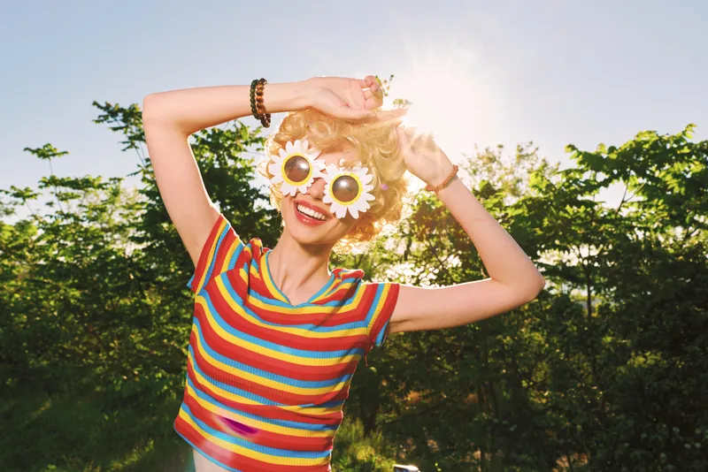 Cheerful woman outdoors, wearing a striped shirt and sunflower sunglasses, basking in warm sunshine, carefree pose, vibrant colors, lush greenery background, joyful spontaneous moment, lively