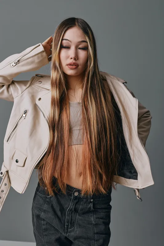 Young woman with long straight hair wearing light-colored jacket and crop top posing with hand behind head against neutral background in studio portrait.