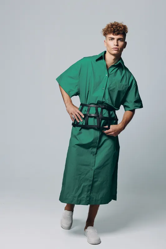 Young man with curly hair wearing green oversized shirt dress and black belt posing in studio with white shoes on light background. Fashion, style and casual outfit concept.