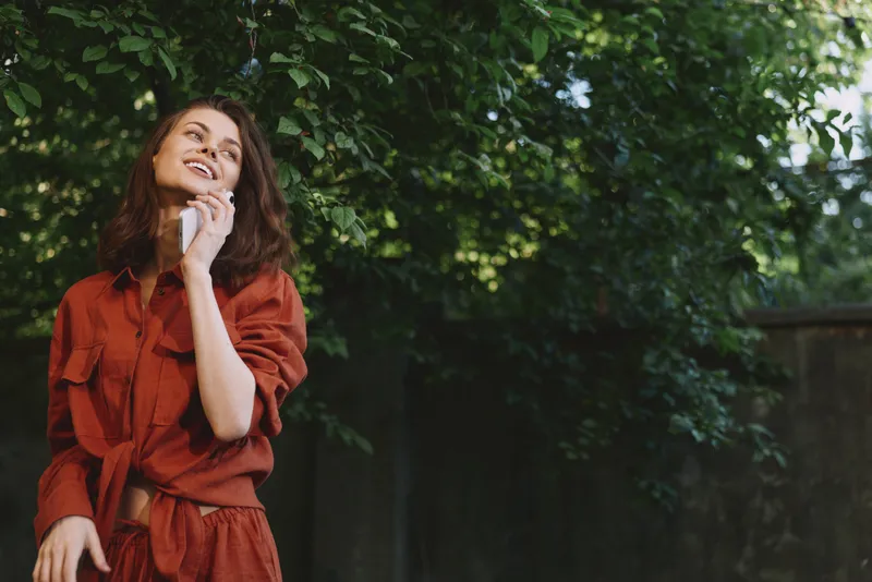 Cheerful woman outdoors, wearing an orange dress, talking on a phone, candid lifestyle moment, smiling gaze, warm sunlight, relaxed pose, lush greenery backdrop, soft breeze, natural colors