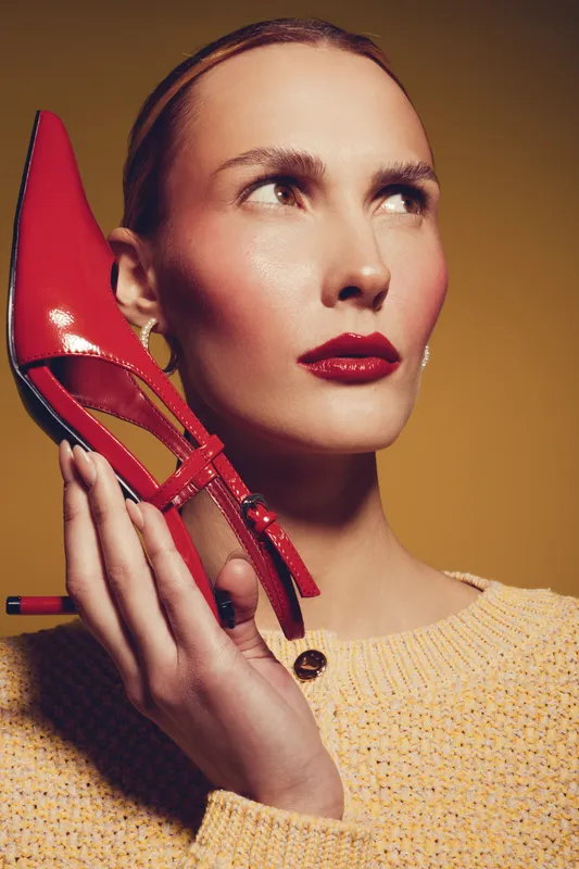 Stylish woman with red lipstick holds red high heel shoe near face, wearing yellow sweater. Studio portrait with warm lighting. Concept of fashion, beauty, and accessories.