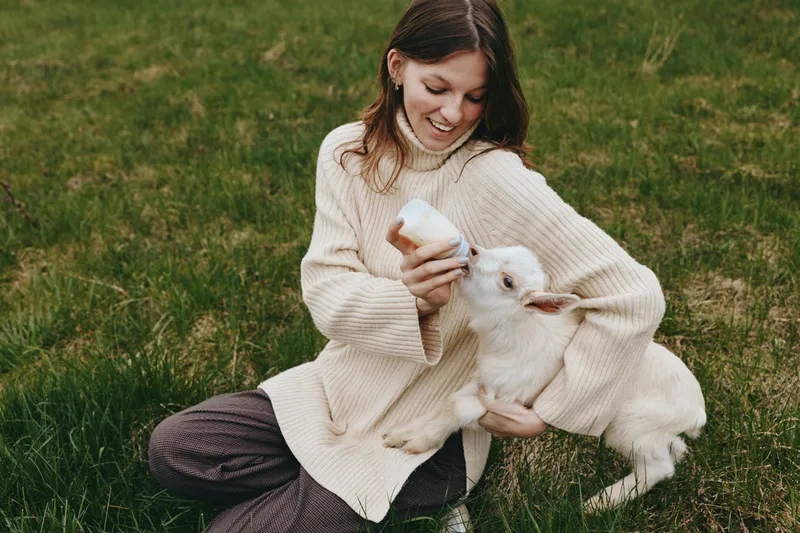A woman in a chunky sweater sits in a grassy field, cradling a goat kid with a warm smile. A candid lifestyle moment highlighting animal companionship and affection.
