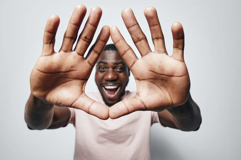 African man with dark skin smiling and showing hands close to camera in heart shape gesture on gray background. Happy male portrait with casual light shirt and joyful expression.