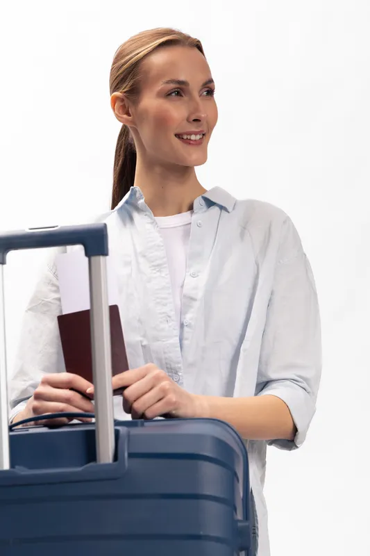 Smiling woman with ponytail holds passport and blue suitcase, looking away. Concept of travel, vacation, and preparation for trip with light background and casual white shirt.