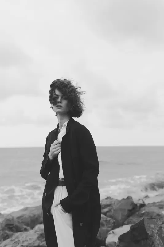 Woman standing on a rocky coastline in a windy seascape, wearing a long dark coat and light trousers, monochrome mood capturing solitude and stormy atmosphere